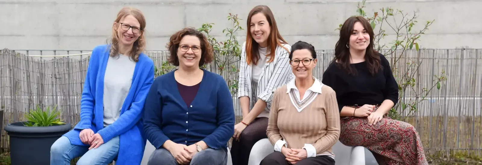 Cinq femmes souriantes sont assises côte à côte sur des chaises modernes en extérieur, sur une terrasse.