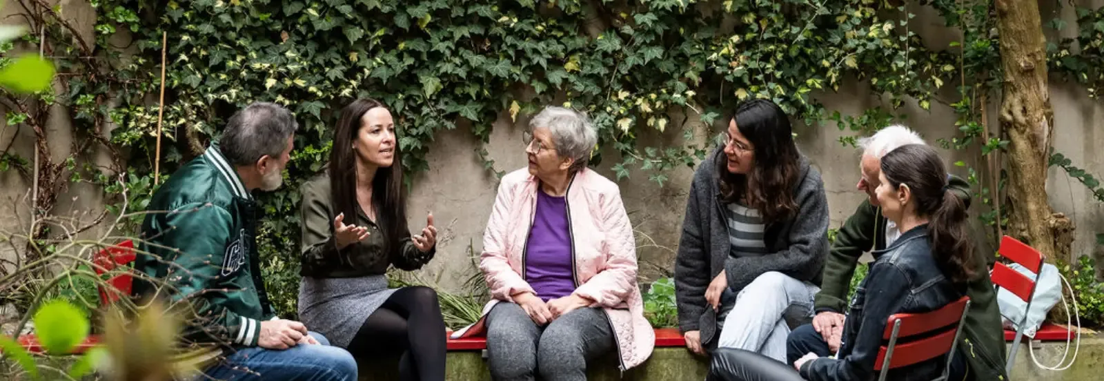 Un groupe de six personnes de différents âges est assis en cercle dans un jardin verdoyant, écoutant attentivement une femme qui s'exprime avec animation.